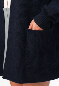 Main dans la poche d'un cardigan en tricot côtelé bleu marine foncé, porté sur une jupe grise et un haut blanc, montrant la texture du tissu et les détails du vêtement.