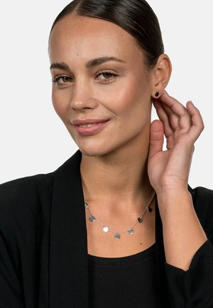 Silberne Halskette mit runden Disc-Anhänger, getragen über einem schwarzen Oberteil. Auch kleine schwarze Steckerohrringe vorhanden. Subtiler Glanz und zeitgenössisches Design.