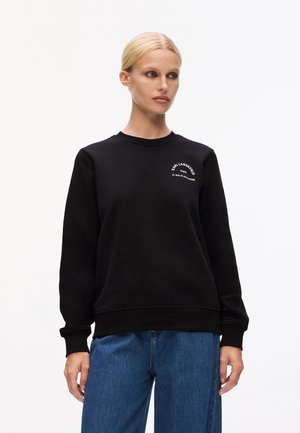Felpa nera con scollo tondo e maniche lunghe. Presenta del testo stampato bianco sul petto sinistro. Abbinata a jeans azzurri chiari.