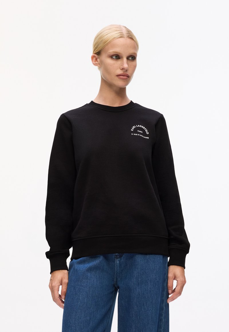 Čierny sweatshir s okrúhlym výstrihom a dlhými rukávmi. Na ľavej hrudi má biele potlačené písmeno. Kombinovaný s svetlomodrými džínsy.