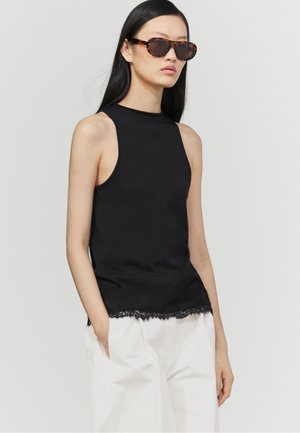 Donna che indossa un top nero senza maniche con orlo in pizzo, pantaloni bianchi e occhiali da sole tartaruga, in piedi davanti a uno sfondo chiaro e semplice.