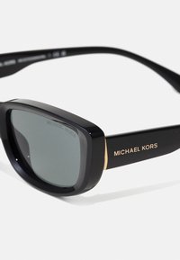 Juodos didelės saulės akinių su pilkais lęšiais, turinčiais lygią plastikinę rėmą ir auksinius akcentus. "Michael Kors" logotipas įspaustas šone.