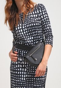 Robe à motifs noir et blanc avec des manches trois-quarts, associée à un sac bandoulière noir avec fermeture éclair et matériel minimaliste.