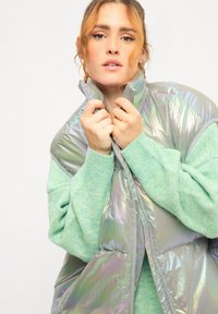 Iridescente opblaasvest met ritssluiting, gelaagd over een zachte, lichtgroene, textuurtrui. Beide kledingstukken hebben een relaxte pasvorm en gedurfde kleuren.