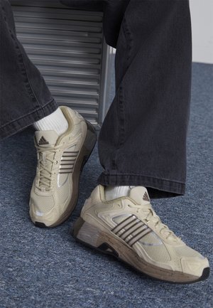 Beige athletic shoes feature mesh material, reflective accents, and a sturdy base with a rubber sole. Worn with dark jeans and white socks.