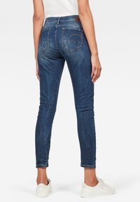 Skinny dark blue jeans made of denim with a mid-rise waist, featuring back pockets and a subtle faded texture. Worn with white sneakers.