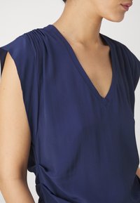 Top blu navy con scollatura a V, in morbido tessuto drappeggiato e dettagli arricciati sulle spalle. Texture liscia con maniche corte.