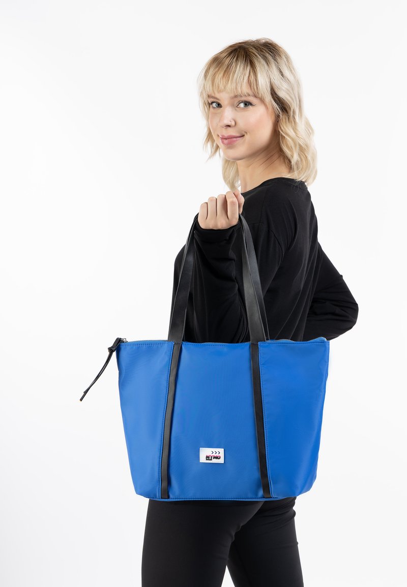 Blå tote bag lavet af holdbart materiale, med sorte håndtag, lynlås lukning og en lille logo lap på forsiden.