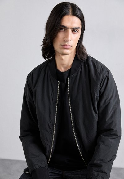 Schwarze Bomberjacke mit einem Reißverschluss vorne, geripptem Kragen und Bündchen, aus glattem Stoff gefertigt, kombiniert mit einem schwarzen Hemd darunter.