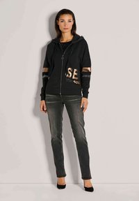 Schwarze Zip-Hoodie mit metallischen Buchstaben, Fronttaschen und Mesh-Details. Kombiniert mit dunklen Jeans und schwarzen High Heels.