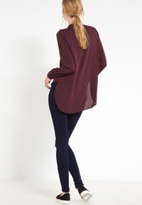 Blusa burdeos de manga larga con un dobladillo curvado, combinada con leggins azul marino. La blusa presenta detalles en los hombros y un panel trasero translúcido.