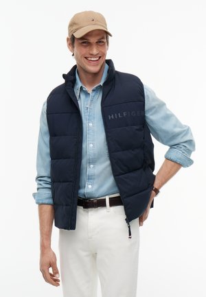 Uśmiechnięty mężczyzna w beżowej czapce, granatowej pikowanej kamizelce z napisem "HILFIGER", jasnoniebieskiej koszuli jeansowej, białych spodniach i brązowym pasku, stojący z jedną ręką na biodrze.