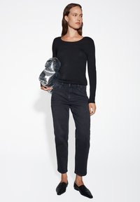 Schwarzes Langarmoberteil, dunkle Jeans und schwarze spitze Loafers; das Modell hält eine silberne, zerknitterte Clutch-Tasche mit einer glatten Oberfläche.