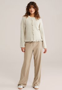 Cardigan tricotat bej deschis, cu tiv cu volane și nasturi, asortat cu pantaloni bej cu dungi subțiri și adidași albi.