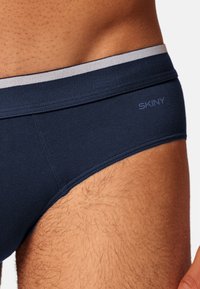 Tummansinisiä alusvaatteita, jotka on valmistettu pehmeästä kankaasta. Niissä on harmaa elastinen vyötärönauha, jossa on painettuna brändin "SKINY" nimi. Lähikuva sivupurkista.