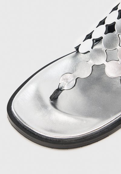 Hopeata metallinen litteä sandaali, jossa on koristeellinen leikattu muotoilu hihnassa, mitä koristaa varvastuki ja musta litteä pohja.