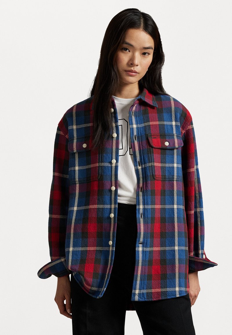 Plaid flannel shirt in red, blue, and black. Button-down design with two chest pockets and rolled sleeves, featuring a textured fabric.