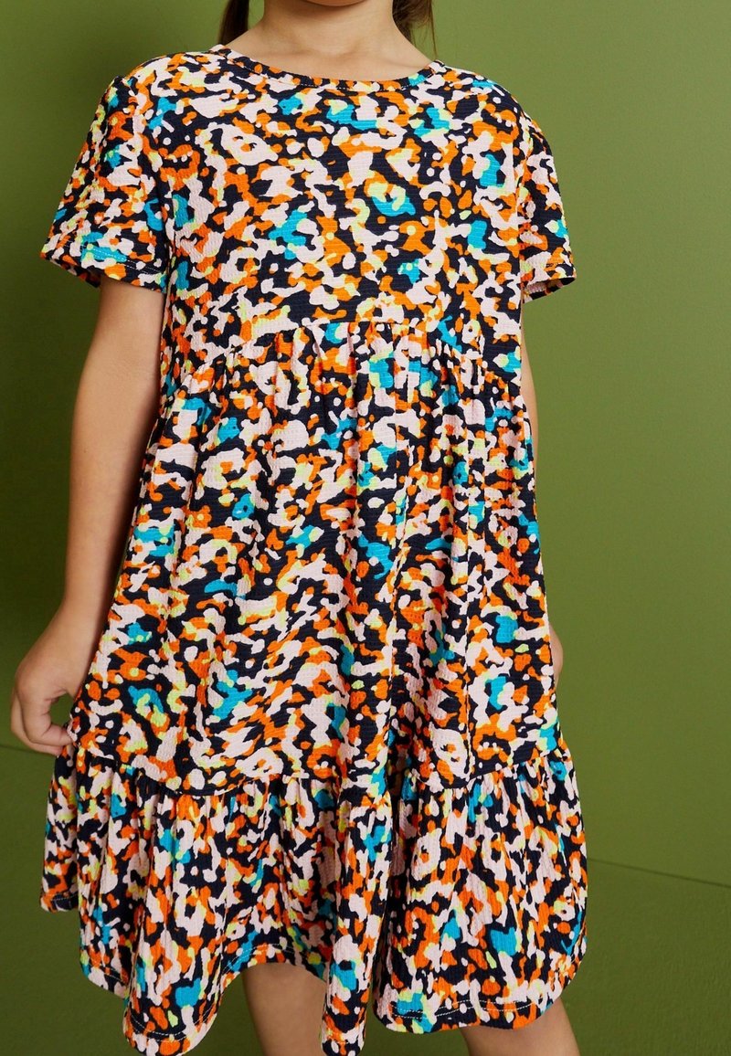 Enfant portant une robe à manches courtes imprimée léopard multicolore avec une jupe froncée sur un fond vert uni.