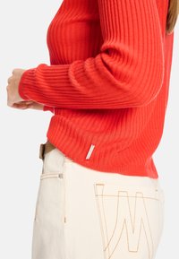 Pull rouge côtelé à manches longues, avec une petite étiquette de marque. Associé à un jean blanc, mettant en évidence des détails de couture contrastants.