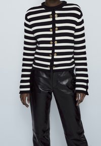 Cardigan a righe bianche e nere con scollo rotondo, caratterizzato da bottoni dorati e texture a coste. Abbinato a pantaloni neri lucidi.