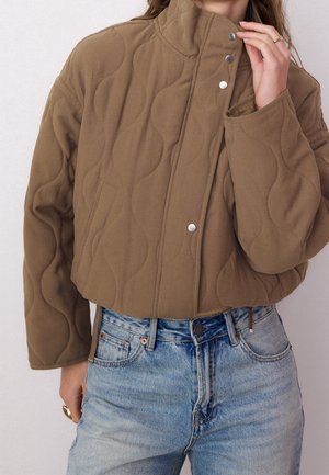 Vrouw draagt een cropped, doorgestikte bruine jas met drukknopen en blauwe high-waisted jeans, waarbij ze met één hand de kraag verstelt.