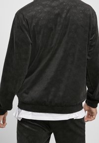 Svart velourjacka med ett texturerat vågmönster. Har elastiska ärmslut och midja, långa ärmar och en avslappnad passform.