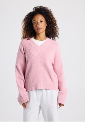 Pink, knitted sweater with a V-neck, ribbed texture, wide sleeves, and a relaxed fit. Paired with light gray sweatpants for contrast.