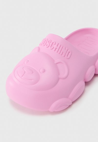 Sabot en caoutchouc rose avec design de visage d'ours, surface douce texturée, et bride de marque embossée "Moschino". Forme arrondie, style décontracté.