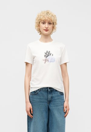 Persoană cu păr blond scurt și creț, purtând un tricou alb cu imprimeu de stea de mare, coral și scoici, asortat cu blugi albaștri.