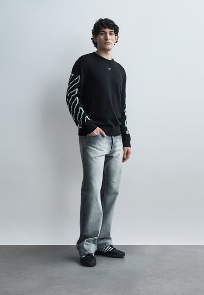 Νεαρός άνδρας στέκεται μπροστά σε γκρίζο τοίχο φορώντας μαύρο πουλόβερ με ανοιχτόχρωμα σχέδια στα μανίκια, ξεβαμμένο γκρι τζιν και μαύρα αθλητικά παπούτσια με λευκές ρίγες.