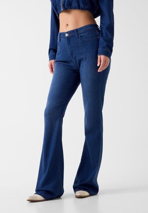 Femme portant un jean flare bleu foncé taille haute, une veste courte bleue et des chaussures beige clair, debout devant un fond blanc uni.