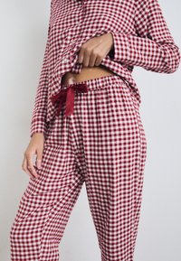 Rød- og hvidternet pyjamas sæt lavet af blødt stof. Langærmet skjorte med knapper og en løs pasform; bukserne har elastik i taljen og en sløjfe.