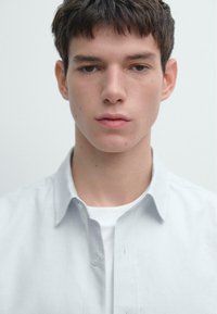 Camicia azzurro chiaro con bottoni, con una trama sottile, indossata sopra una t-shirt bianca. Tessuto liscio, colletto a punta e bottoni visibili. Sfondo trasparente.