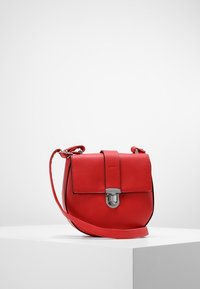 Červená kožená crossbody taška se stříbrným zapínáním na bílém podstavci proti čistému bílému pozadí.