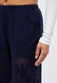 Κοντινό πλάνο στην κάτω κοιλιακή περιοχή και το χέρι ενός ατόμου που φοράει μπλε-σκούρο παντελόνι με λουλουδάτη δαντέλα και άσπρο μακρυμάνικο τοπ, με φόντο απλό.