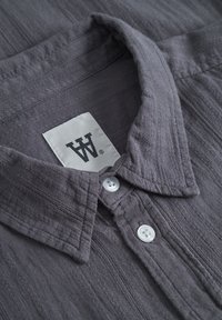 Gray textured shirt featuring a collared neckline, two white buttons, and a tag with a logo on a plain background.