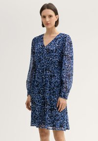 Blau gemustertes Kleid mit V-Ausschnitt, langen transparenten Ärmeln und gerafftem Taillenbereich. Der Stoff weist eine Mischung aus dunklen und hellen Blautönen auf.
