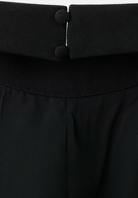 Crni top s drapiranim dizajnom, izrađen od glatkog materijala. Uočljive okrugle dugmadi na izrezu i uredno šiven rub.