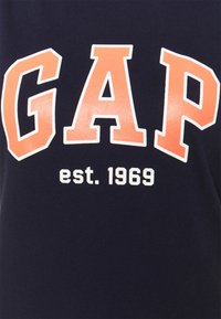 Námořnická modrá bavlněná tričko s velkým nápisem "GAP" v oranžových písmenech a "est. 1969" menším bílým fontem pod ním. Měkká textura.