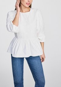 Biała bawełniana bluzka z bufkami i peplum, z teksturowanymi wzorami. Połączona z niebieskimi dżinsami.