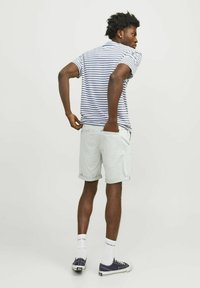 Polo w paski w kolorze białym i granatowym, jasnoszare szorty, podwinięte mankiety, casualowe sneakersy, postawa stojąca z rękami w kieszeniach, neutralne tło.