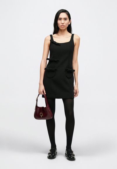 Fekete ujjatlan ruha masni díszítéssel, két nagy zsebbel és testhez simuló szabással. A modell bordó táskát tart, fekete harisnyát és cipőt visel.