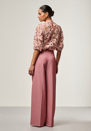 Vrouw staand, gekleed in roze wijde broek en een bloemen kantblouse met pofmouwen, zijwaarts staand tegen een effen achtergrond.