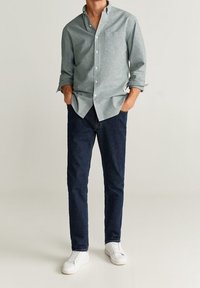 Camisa de botones gris claro con un bolsillo en el pecho, combinada con jeans azul oscuro y zapatillas blancas. La camisa presenta mangas enrolladas.