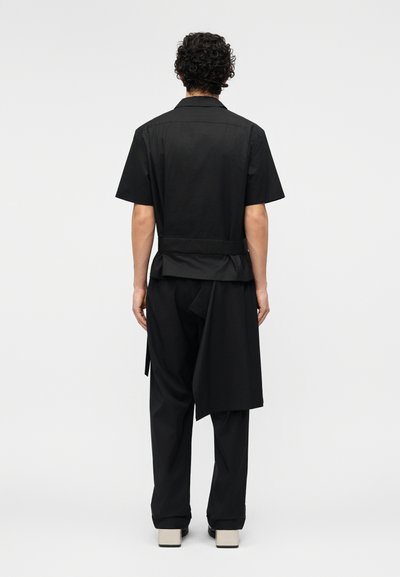 Άνδρας με σγουρά μαλλιά στέκεται γυρισμένος προς τα πίσω, φορώντας κοντομάνικο μαύρο πουκάμισο, μαύρο φαρδύ παντελόνι και μπεζ παπούτσια με τετράγωνη μύτη σε απλό φόντο.