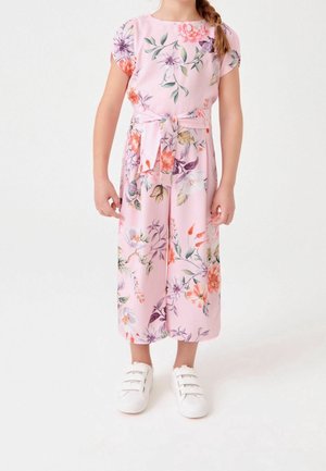 Enfant portant une combinaison à fleurs rose avec une ceinture nouée à la taille et des baskets blanches à scratch, debout sur un fond blanc uni.