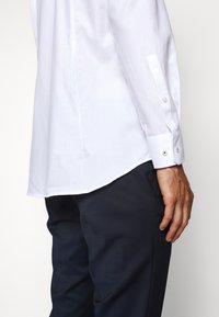 Bílá košile s dlouhým rukávem, zakřiveným lemem, zapínáním na manžety a texturovanou látkou, doplněná tmavě modrými kalhotami.