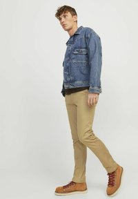 Denimtakki, jossa on kaksi etutaskua, beige istuvat housut ja ruskeat vaelluskengät punaisilla nauhoilla. Neutraali tausta ilman häiriötekijöitä.
