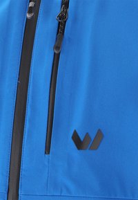 Sininen synteettinen takki, jossa on sileä pinta, mustat vetskarikoristeet ja erottuva musta logo oikealla puolella.