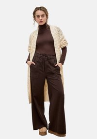 Lange crèmekleurige gebreide cardigan over een bruin coltrui-top en donkerbruine wijde broek. Model draagt lichttan platform schoenen.